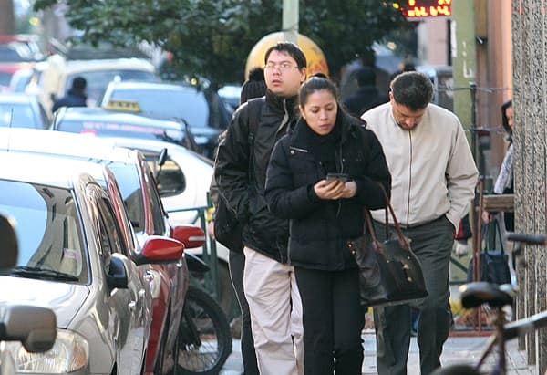 Tres personas caminan por una vereda en una calle céntrica de Asunción, vistiendo camperas gruesas. Una mujer observa su celular mientras un hombre a su lado camina con las manos en los bolsillos. Otro hombre, detrás, lleva una mochila y mira al frente. El entorno urbano muestra vehículos estacionados, bicicletas y un fondo con árboles y más transeúntes, evidenciando una mañana fría y activa.