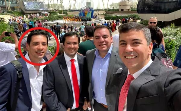 Cuatro hombres vestidos formalmente posan para una selfie en un evento internacional al aire libre, con una multitud y un escenario colorido al fondo. El hombre ubicado a la izquierda de la imagen, señalado con un círculo rojo, destaca por ser el protagonista del contenido relacionado con un nombramiento político. La escena ocurre durante el día, en un ambiente soleado y moderno, con arquitectura contemporánea y presencia de delegaciones internacionales.