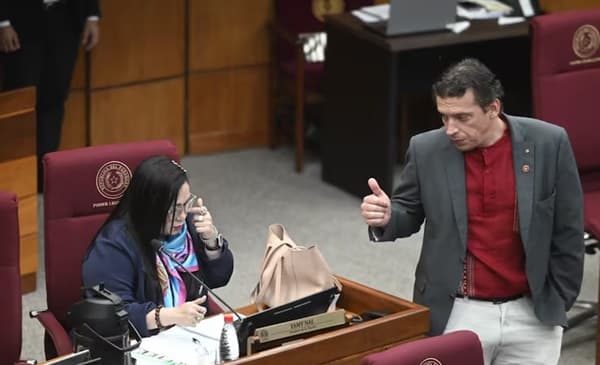 En la imagen se observa a dos senadores paraguayos en el recinto del Senado Nacional. A la izquierda, la senadora Norma Aquino se encuentra sentada en su banca senatorial, sosteniendo un teléfono celular cerca de su oído mientras mantiene una conversación. A la derecha, el senador Juan Carlos Galaverna está de pie junto a su asiento, vestido con un saco gris y camisa roja, haciendo un gesto con su mano derecha levantada mientras parece dirigirse hacia su colega. El ambiente corresponde al hemiciclo del Congreso Nacional, con las características bancas de color bordó y el mobiliario institucional típico del recinto legislativo paraguayo.